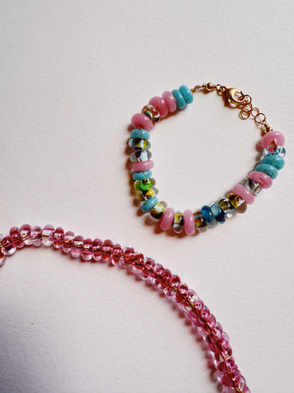 The Donut Bracelet, featuring pink, blue, and multicolored beads with a gold-filled clasp, is displayed beside a string of translucent pink beads on a white surface.