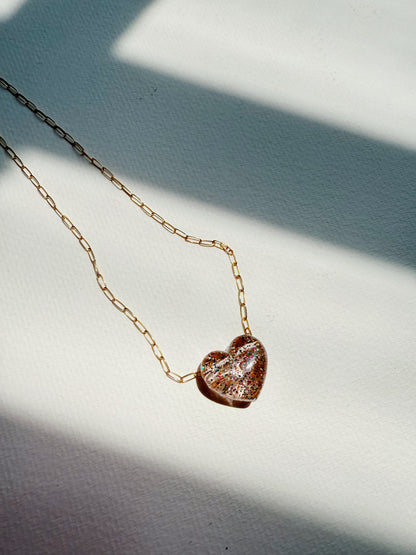 The Acrylic Heart Necklace, featuring a glittery acrylic heart pendant on a 14k gold filled chain, rests on a textured white surface in sunlight, casting soft shadows.