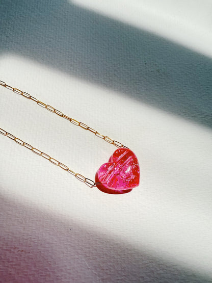 The Acrylic Heart Necklace features a gold filled chain and a translucent pink acrylic heart pendant, displayed on a textured white surface with soft sunlight and gentle shadows.