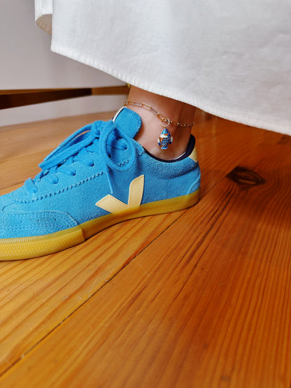 A person wearing bright blue sneakers and a white garment stands on a wooden floor, showcasing the Fish Anklet—a gold-plated chain with a small blue and red cloisonné fish charm.