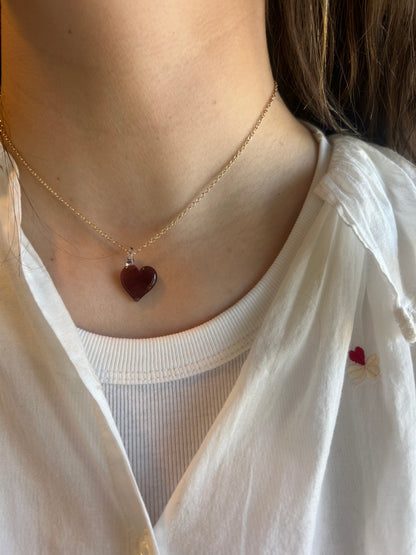 A person wearing the Flat Glass Heart Layer—a 14k gold chain necklace with a small red handblown glass heart pendant—styled over a white ribbed top and an embroidered white shirt featuring a tiny red heart near the collar.