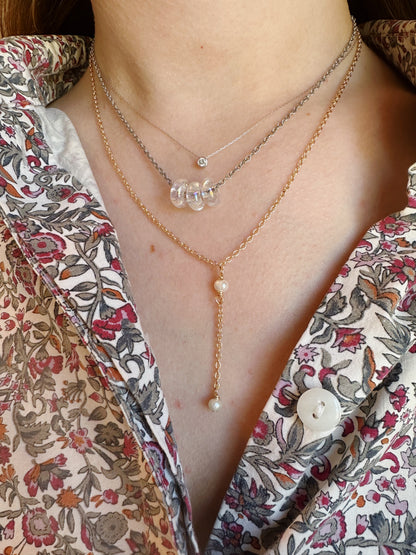 A person wears a floral shirt and layered necklaces, including the Double Pearl Lariat—a 14k gold filled chain with cultured freshwater pearls and a vertical drop—plus a clear bead strand and one with a small pendant.