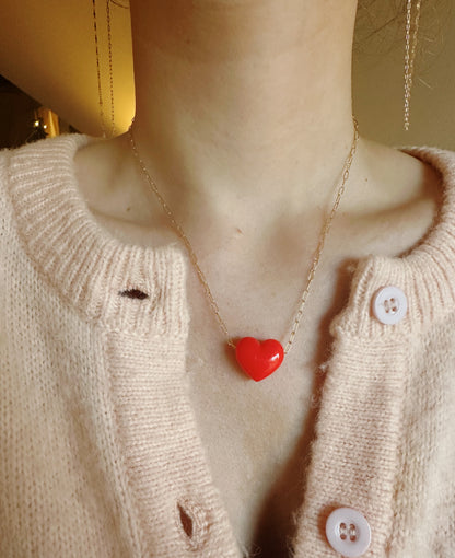 Shown from the shoulders up, a person in a buttoned cream sweater with small holes wears the Acrylic Heart Necklace, featuring an acrylic heart on a 14k gold filled chain that adds elegance to their look.