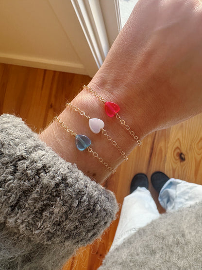 A hand wearing three Friendship Bracelets—14K gold filled chains with blue, white, and red acrylic heart charms. The person wears a gray sweater, light jeans, black shoes, and stands on a wooden floor by a window.