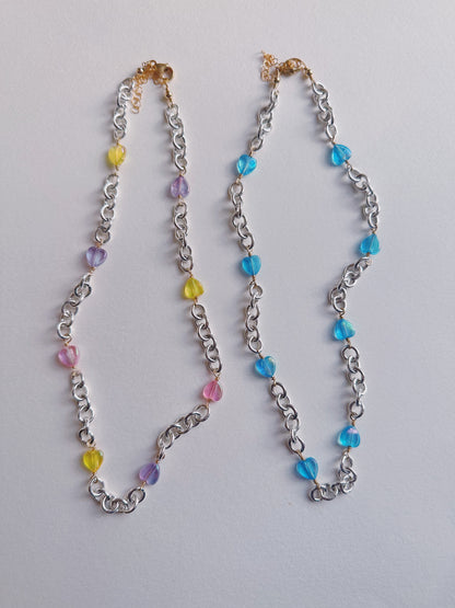 Two Sterling Silver Candy Layer necklaces are shown on a white surface: one with yellow, pink, and purple acrylic hearts; the other with blue beads. Both feature sterling silver chains and 14k gold filled clasps.