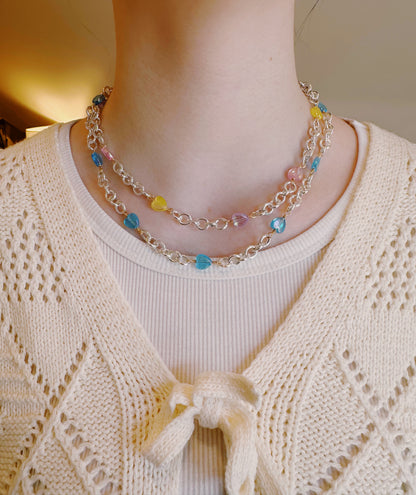 A person wears a cream knit sweater with a bow at the neckline, paired with the Sterling Silver Candy Layer—a chain necklace adorned with small acrylic heart beads in blue, yellow, and pink.