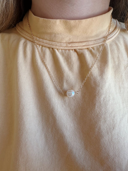 A person wearing a pale yellow high-neck top and the Dainty Single Pearl Layer—a delicate 14k gold filled chain necklace with a single cultured freshwater pearl pendant.