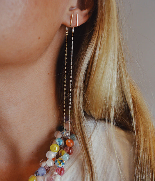 A close-up of a woman in a white top and the KK x Poema collection: colorful beaded necklaces, 14k gold filled chain earrings with minimalist bars, and freshwater pearl accents. Her blond hair falls loosely over her shoulder.