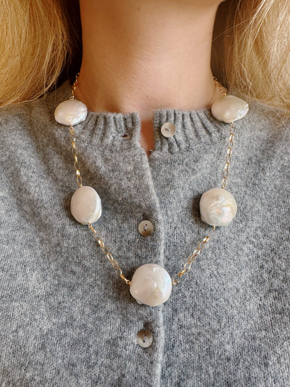 A woman wears a grey sweater and a gold chain necklace called Chunky Pearl Layer, featuring large, flat freshwater pearls and 14k gold filled details.