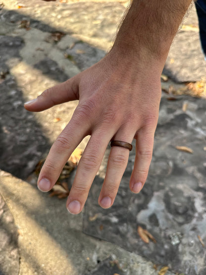 A close-up of a left hand wearing the Wooden Ring- Striped on the ring finger, with a stone surface and scattered dried leaves in the background.
