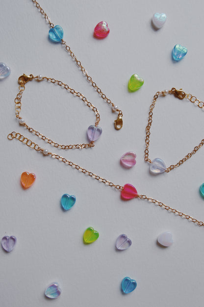 Friendship Bracelets with colorful acrylic heart beads and delicate 14K gold-filled chains are displayed on a light surface, surrounded by scattered small, bright heart beads for a playful touch.