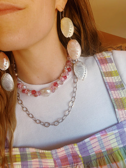 A woman wears Rainbow Abalone Drops earrings made from real shells, layered pink and clear beaded necklaces, and a white top under a pastel plaid dress. Only the lower half of her face and her long hair are visible.