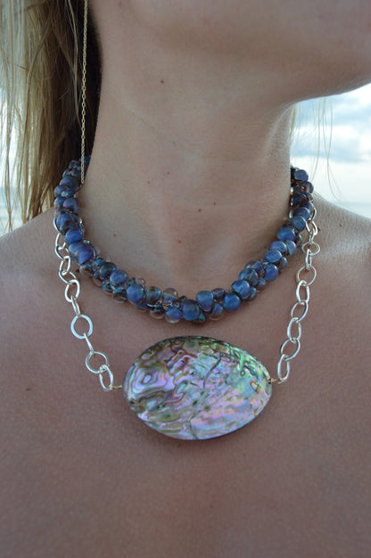 A close-up shows a woman’s neck wearing the Sterling Silver Abalone Layer: two necklaces—one with blue stones, the other with a large oval abalone shell pendant on a sterling silver chain. Pale skin and part of her shoulder are visible.