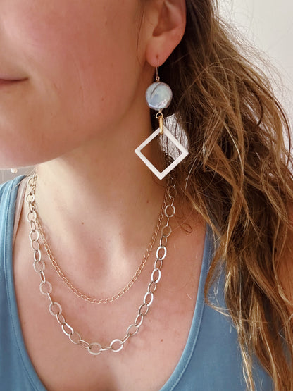 A person with wavy, light brown hair wears a blue top, a layered gold chain necklace, and Diamond Shell and Pearl Drops earrings featuring a round blue stone and a dangling white diamond-shaped shell.