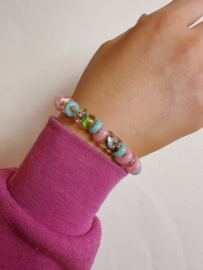 A hand in a pink long-sleeve top displays the Donut Bracelet, featuring colorful handblown glass beads and gold filled findings, against a plain light background.