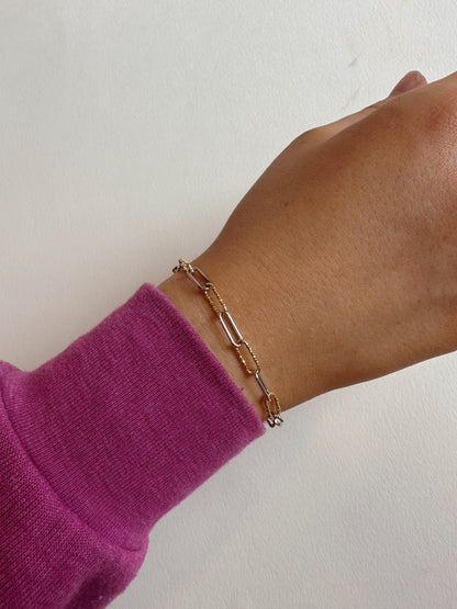 A hand wearing the Mixed Metal Glitz Bracelet, featuring a gold filled chain with oval links, is shown against a white background. The person has on a long-sleeve purple top.