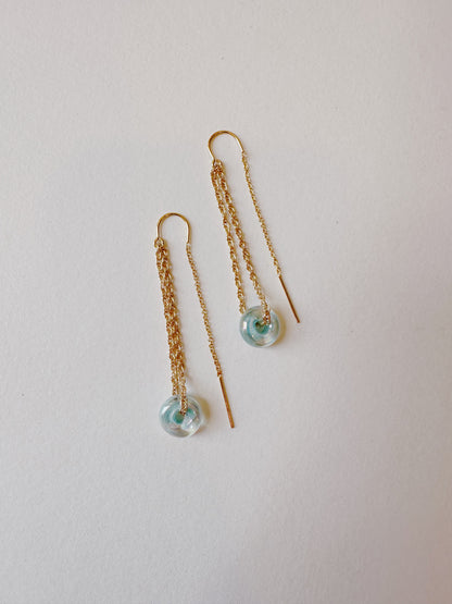 The Glass Donut Threaders are 14k gold filled earrings with delicate chains and small, round, light blue handblown glass beads at the ends, shown on a plain white background.
