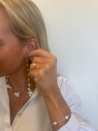 A blonde woman in a white shirt holds a long pearl earring to her ear. She wears the Pearl Bracelet with cultured freshwater pearls, a matching necklace, and a 14k gold filled ring with a square gemstone against a plain background.