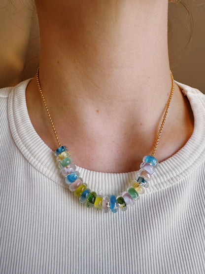 Wearing a white ribbed shirt, a person showcases the Lots of Donuts Layer—a 14k gold filled chain necklace with vibrant handblown glass beads in blue, green, yellow, and clear—resting just above the collarbone.
