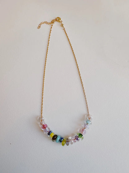 The Lots of Donuts Layer necklace showcases handblown glass beads in clear, green, blue, yellow, and pink on the lower part of a 14k gold filled chain, presented against a plain white background.
