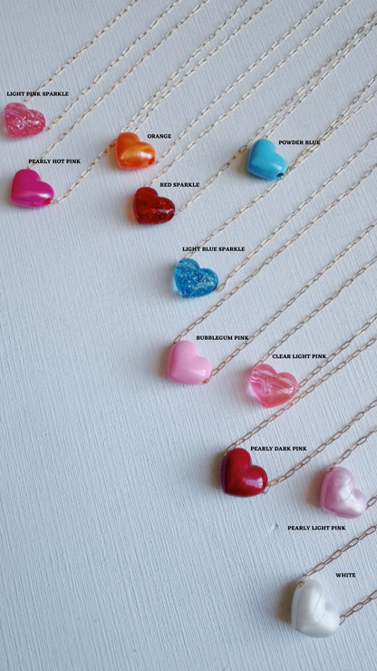 Nine Acrylic Heart Necklaces in assorted hues—light pink sparkle, pearly hot pink, orange, red sparkle, blue tones, and more—shine on a light background. Each acrylic heart is strung on a 14k gold filled chain for lasting elegance.