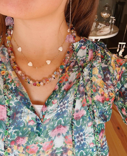 A woman wearing a colorful floral top and the Mixed Metal Mother of Pearl Layer necklace stands in a bright room with jewelry displayed in the background.