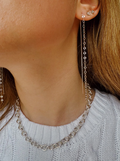 A close-up of a woman's neck and ear, featuring Glitzy Threaders long silver chain earrings, a chunky sterling silver chain necklace, and small stud earrings. She wears a white knit sweater.