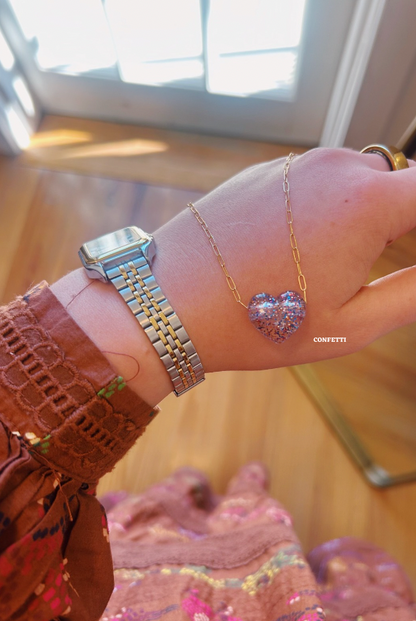 A hand displays a silver watch and a 14k gold filled chain bracelet with a glittery blue Acrylic Heart Necklace; sunlight streaming through a window highlights the elegance of gold filled jewelry.