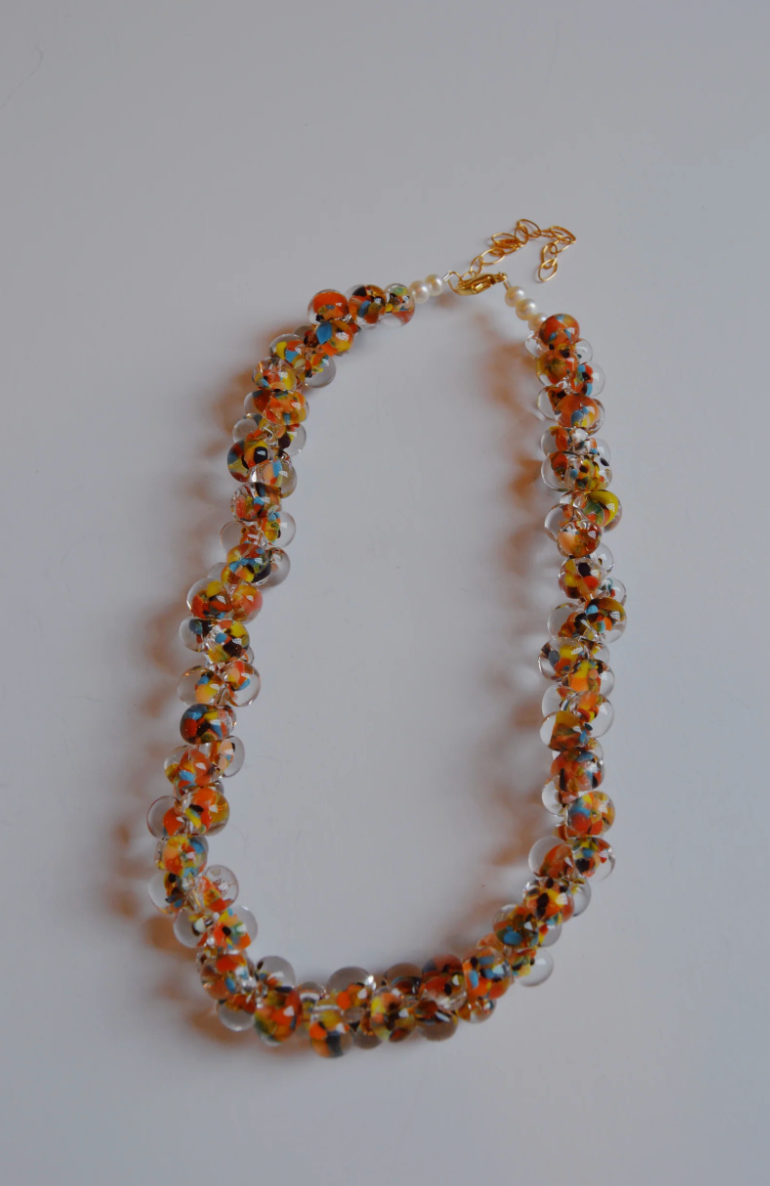 The Print Bubble Glass Necklace showcases clear, orange, yellow, and blue beads in a twisted design, set against a plain white background.