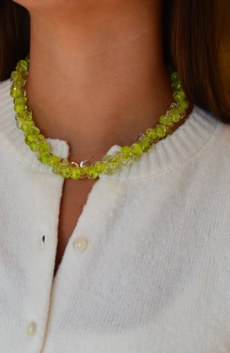 A person wears a white buttoned sweater and the Solid Bubble Glass Necklace, featuring beaded green and clear shimmering glass. The focus is on the necklace’s glimmering beads and the upper part of the cozy sweater.