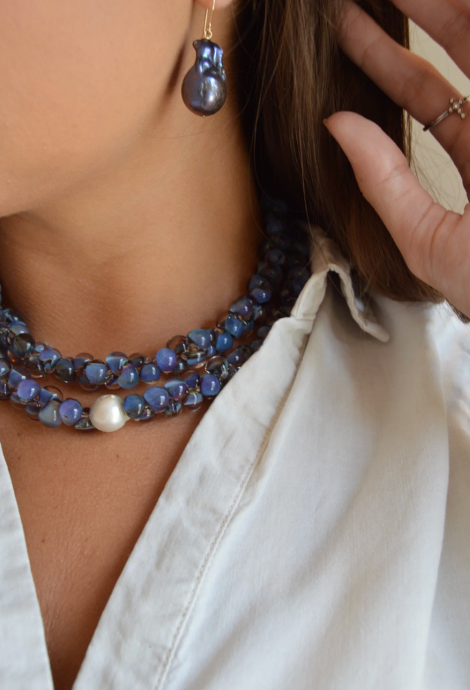 A person wearing a white collared shirt and the Print Bubble Glass Necklace, along with a dark blue dangling earring and a 14k gold filled ring, touches their hair.
