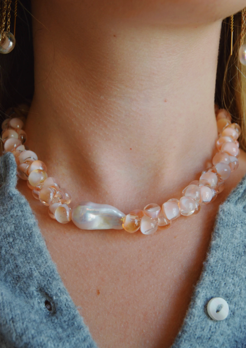 A close-up of a person wearing the Solid Bubble Glass Necklace, featuring pale pink stones, freshwater pearls, and a central irregular white pearl, styled with a light blue textured button-up top.