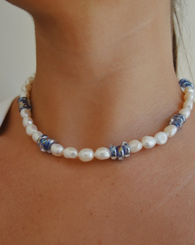 A close-up of someone wearing the Glass Donut and Pearl Layer necklace, featuring freshwater pearls and clusters of small blue glass beads, set against tan skin.