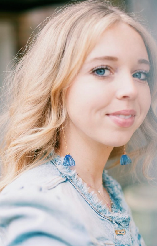 A young woman with long blonde hair and blue eyes, in a light denim jacket, smiles softly at the camera. She wears Glass Tulip Earrings and a delicate 14k gold-filled chain. The softly blurred background highlights her look.