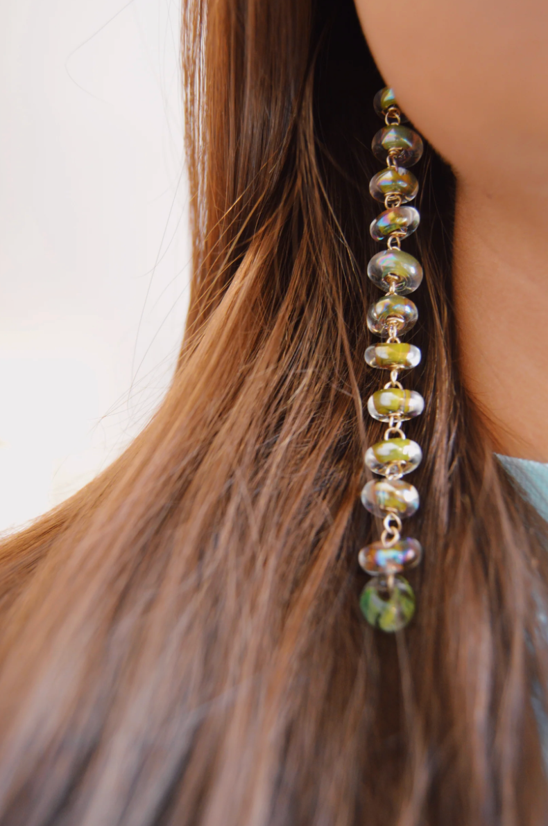 Close-up of a person with long brown hair wearing Donut Drops earrings in Shimmer Olive. Handmade glass beads with gold details stand out against a light, blurred background, highlighting this artisan jewelry piece.