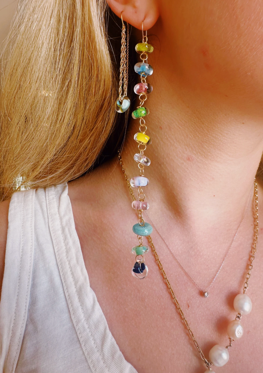 A close-up of a person in a white sleeveless top wearing rainbow Donut Drops earrings, crafted from handmade glass beads, plus layered gold necklaces—one with pearls. Blonde hair partially covers the ear.
