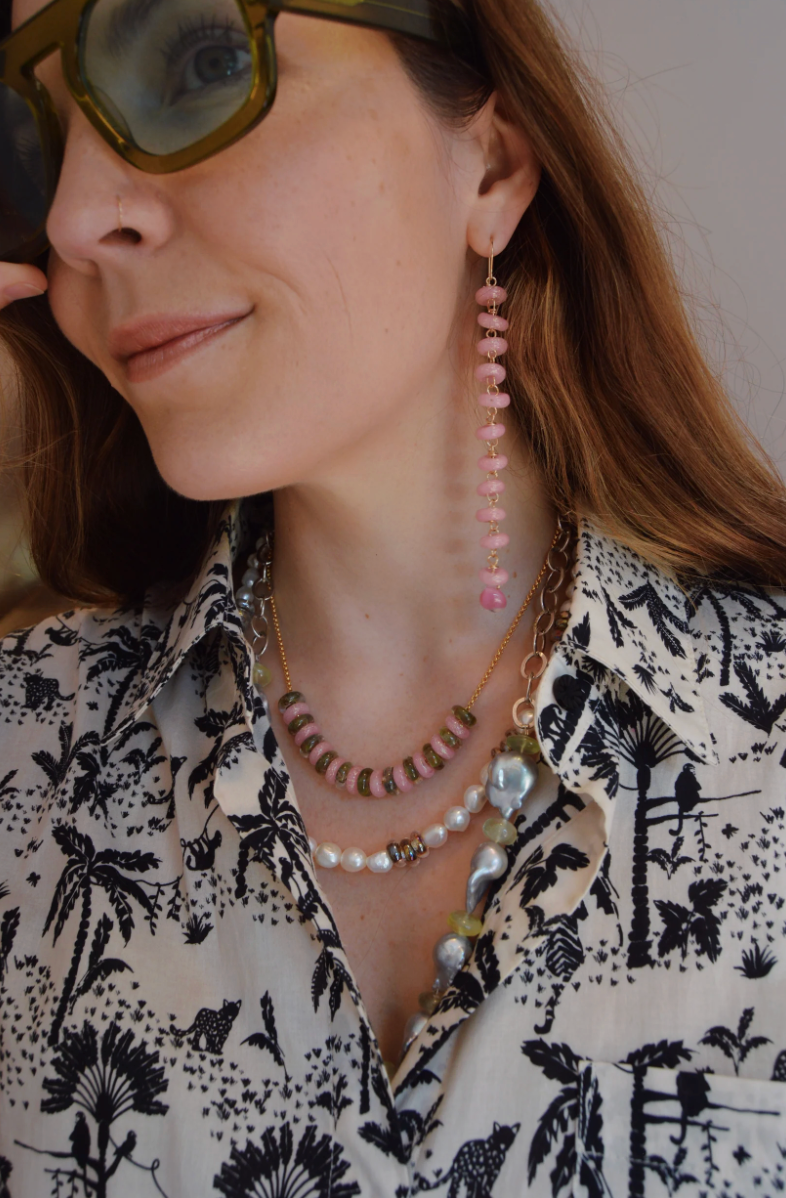 A woman with long brown hair, sunglasses, and a nose ring wears layered necklaces and Donut Drops earrings in Pink Sparkle as she gently touches her sunglasses.