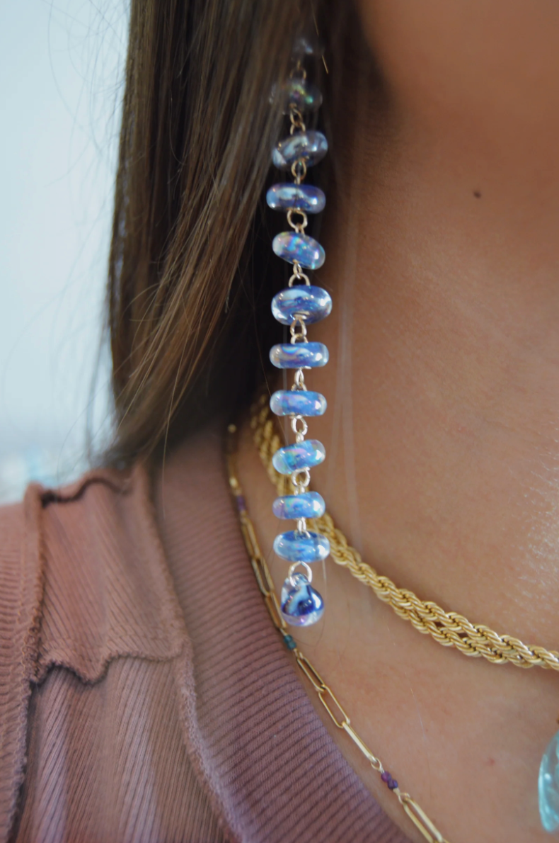 Close-up of a person in a mauve top wearing the Donut Drops earrings in Shimmer Indigo, featuring stacked artisan lampwork beads, along with two gold chain necklaces.