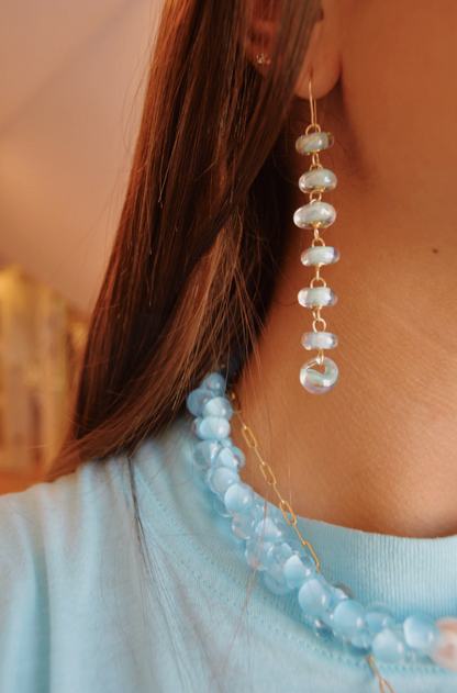 Close-up of a person wearing Donut Drops earrings in Shimmer Blue—stacked translucent lampwork beads—and a matching necklace, with long brown hair and a light blue shirt, perfect for artisan jewelry lovers.
