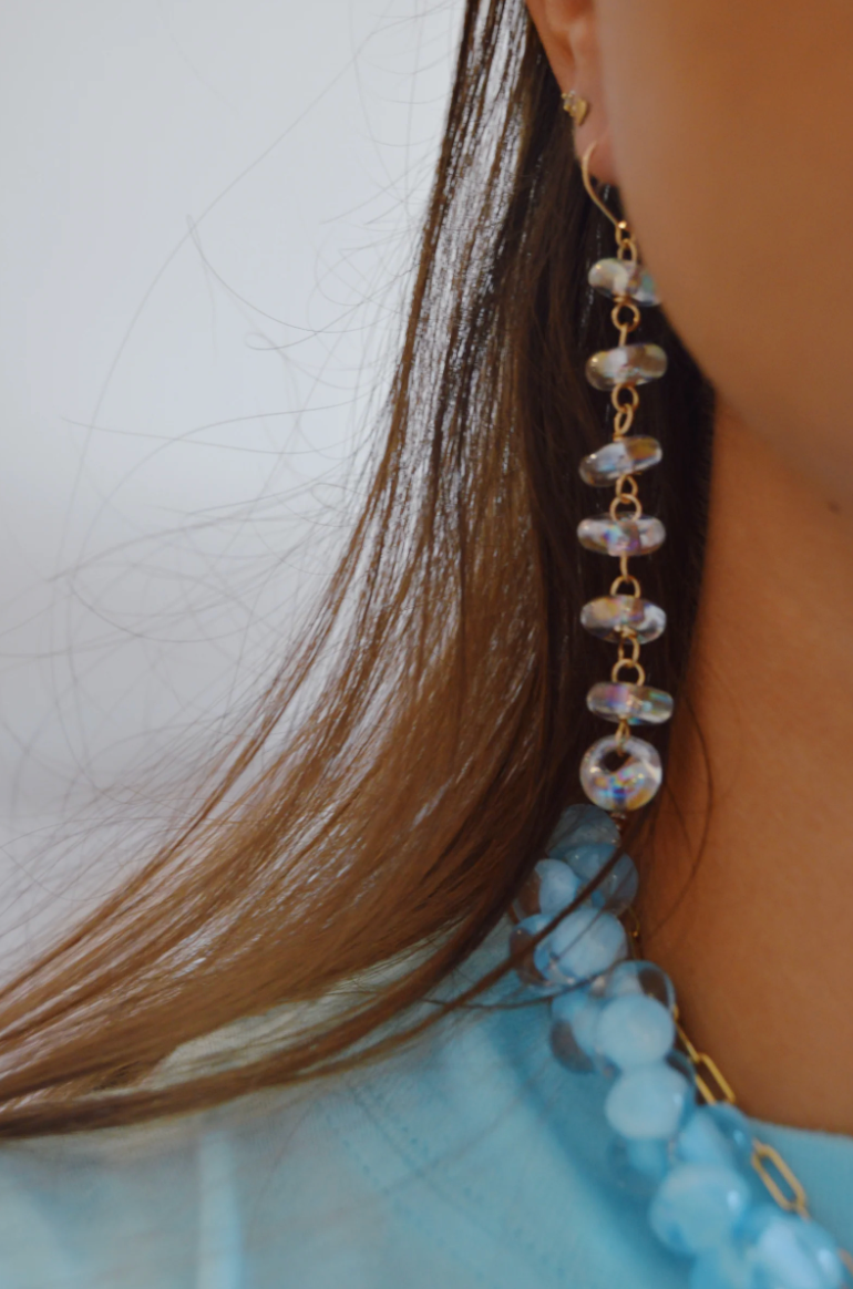 Close-up of a person wearing Donut Drops earrings in Shimmer Clear, crafted from handmade glass beads, paired with a matching blue beaded necklace. They wear a light blue top, with brown hair against a light background.