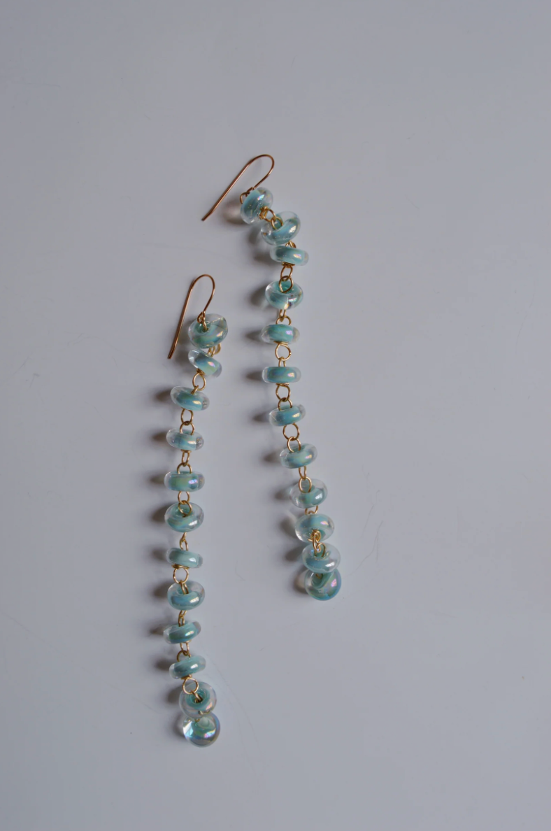 Donut Drops earrings feature shimmer blue handmade glass beads linked vertically on gold hooks, highlighting artisan craftsmanship against a plain white background.