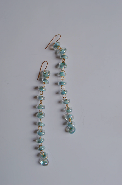 Donut Drops earrings feature shimmer blue handmade glass beads linked vertically on gold hooks, highlighting artisan craftsmanship against a plain white background.