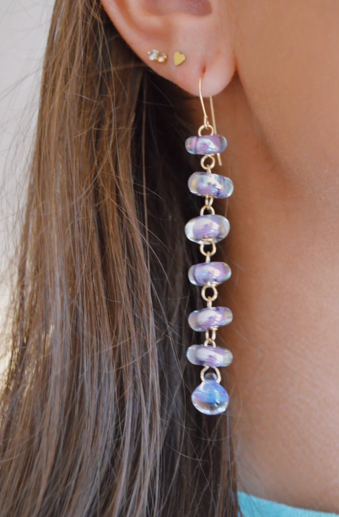 Close-up of a woman's ear with long brown hair, wearing artisan jewelry: Donut Drops earrings in Shimmer Lavender, featuring stacked handmade glass beads linked by gold wire. She also has two small gold studs in her upper ear lobe.