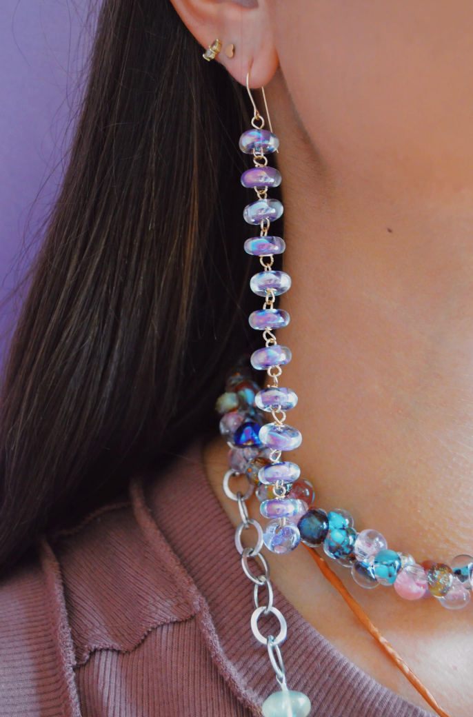 A close-up of a person with long brown hair, wearing Donut Drops earrings and necklace in Shimmer Lavender; the artisan jewelry features iridescent lampwork beads. The person wears a mauve top against a purple background.