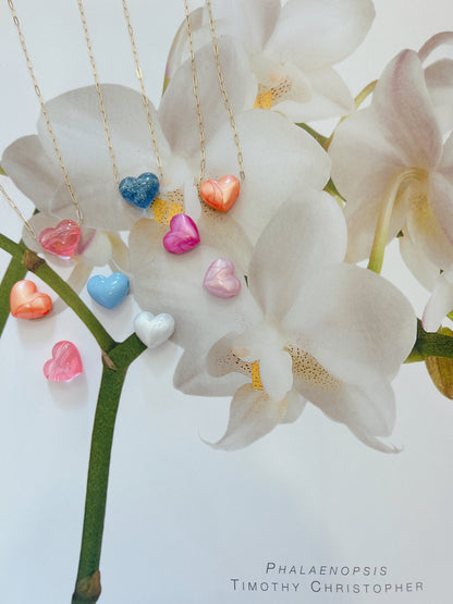 Acrylic Heart Necklaces in various colors hang from delicate 14k gold filled chains, arranged over a photo of white Phalaenopsis orchids with yellow centers. The names Phalaenopsis and Timothy Christopher are shown below.