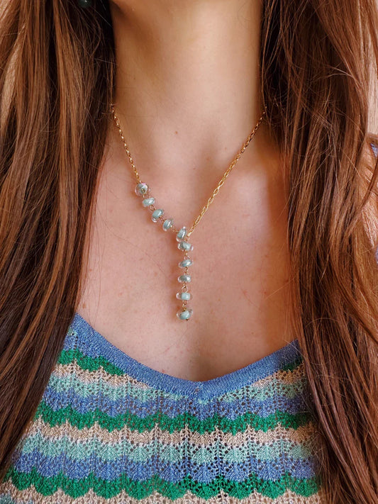 A person with long brown hair wears the Donut Lariat, a 14k gold filled chain necklace featuring blue gemstones in a vertical line, over a blue and green patterned knit top.