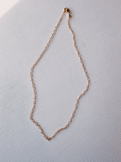 The Heart Chain Layer features a delicate 14k gold filled chain with small, oval links, arranged in an irregular, rounded shape on a textured white surface.