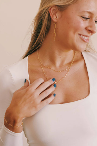 A woman with long blonde hair wears the Heart Chain Layer 14k gold filled necklace, a gold bracelet, drop earrings, and a white square-neck top. She smiles and touches her necklace with her blue-painted nails.