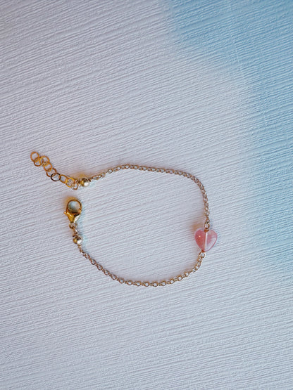 The Gemstone heart bracelet features a delicate 14k gold filled chain with a small cherry quartz heart charm and an adjustable clasp, displayed on a textured white and blue surface. Its simple, elegant design makes it perfect for any occasion.