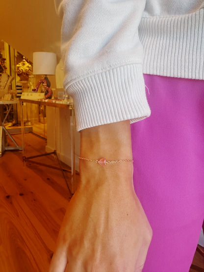 A person in a white sweater and bright pink pants showcases the Gemstone heart bracelet—a delicate 14k gold filled chain with a small heart charm—on their wrist, standing on a wooden floor with a decorated table in the background.